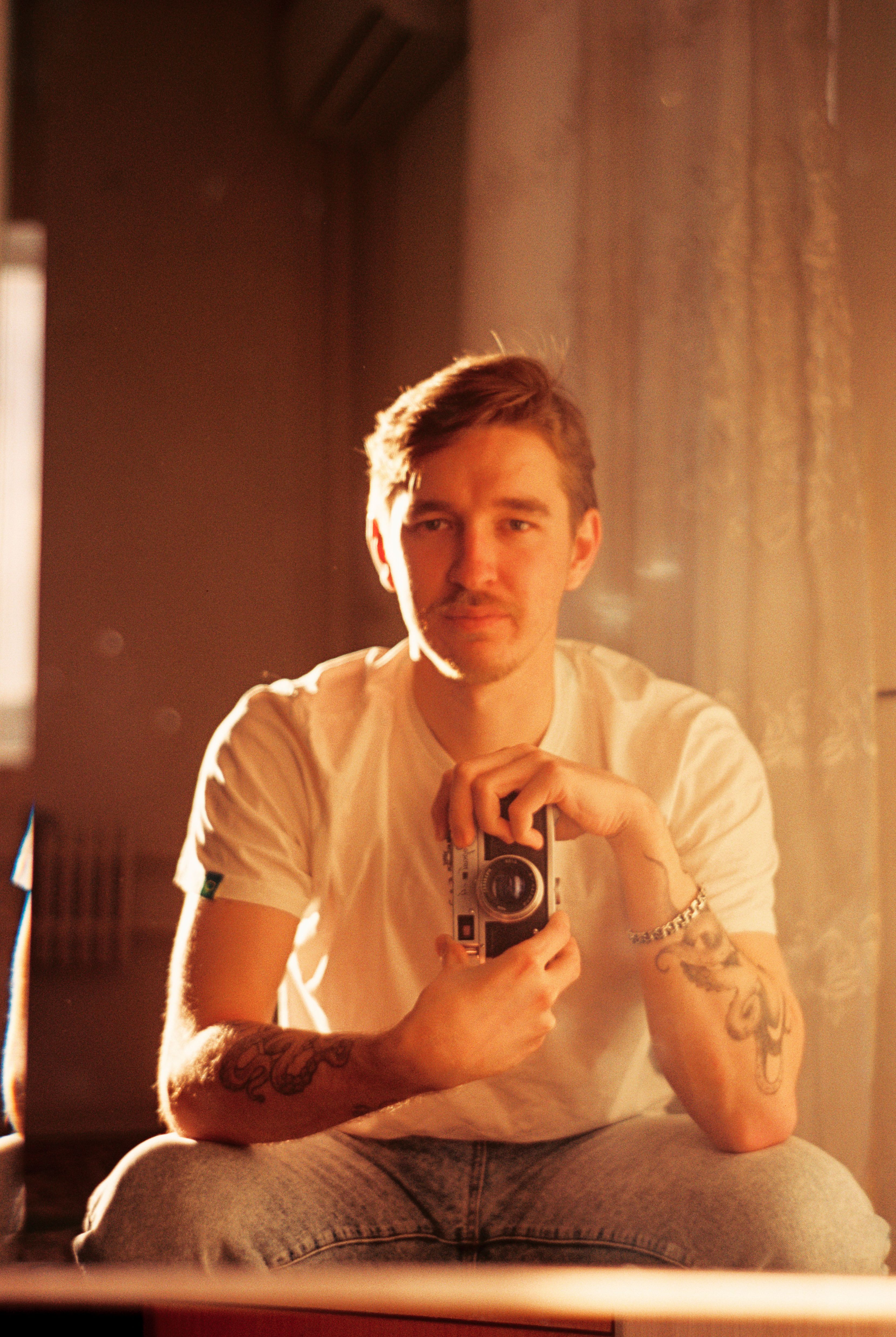 Portrait of a young man with tattoos holding a vintage camera, sitting in a warmly lit room.