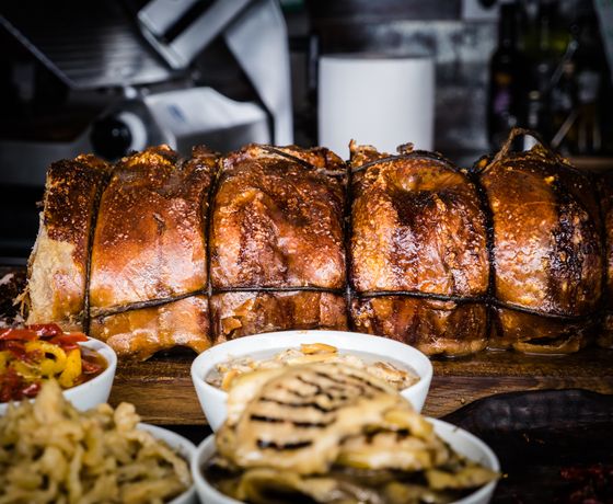Long twine-tied porchetta roast with crackling skin behind bowls of peppers and grilled sides