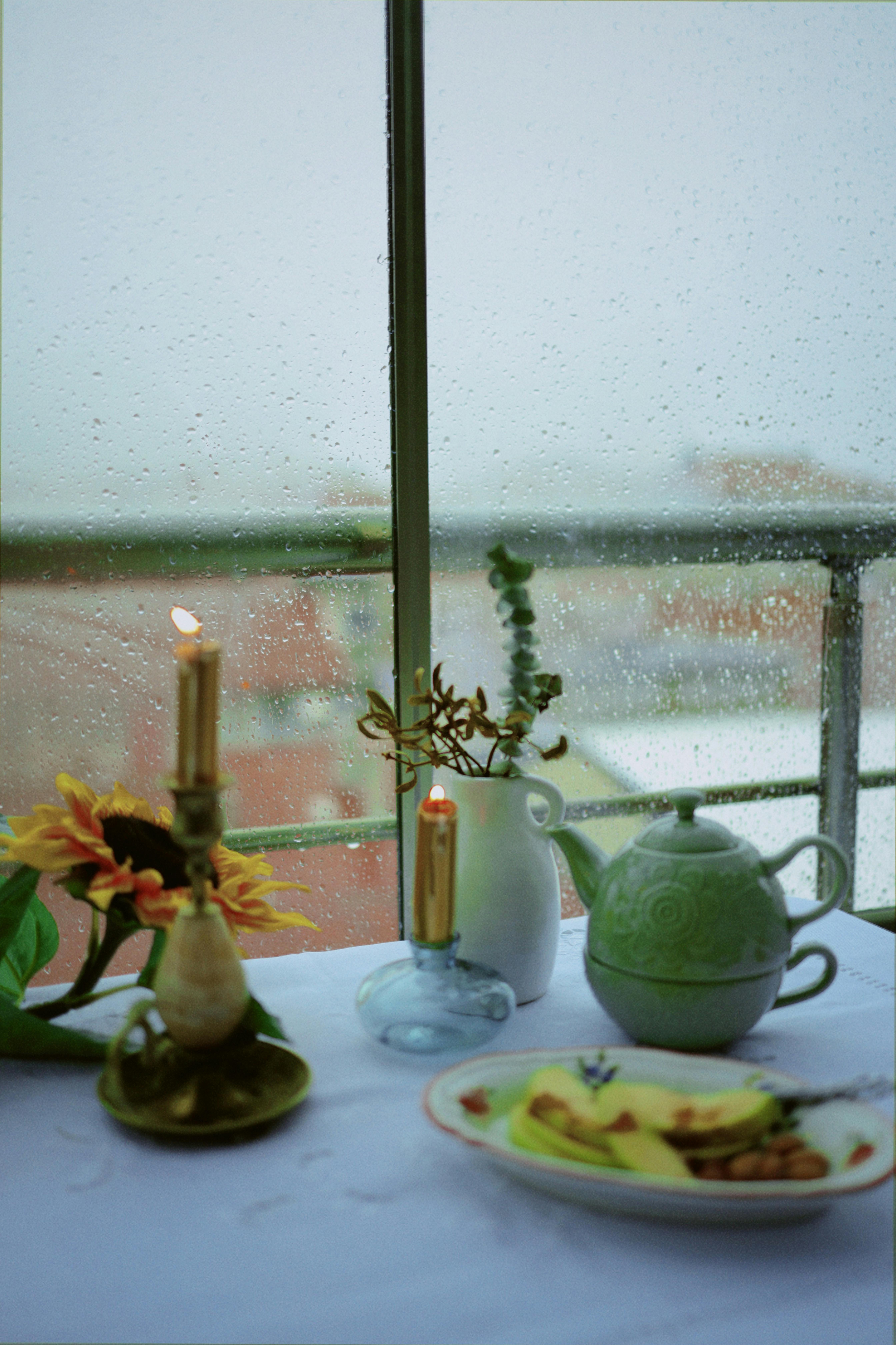 A serene indoor tea setup with candles on a rainy day, featuring a teapot and a window view.