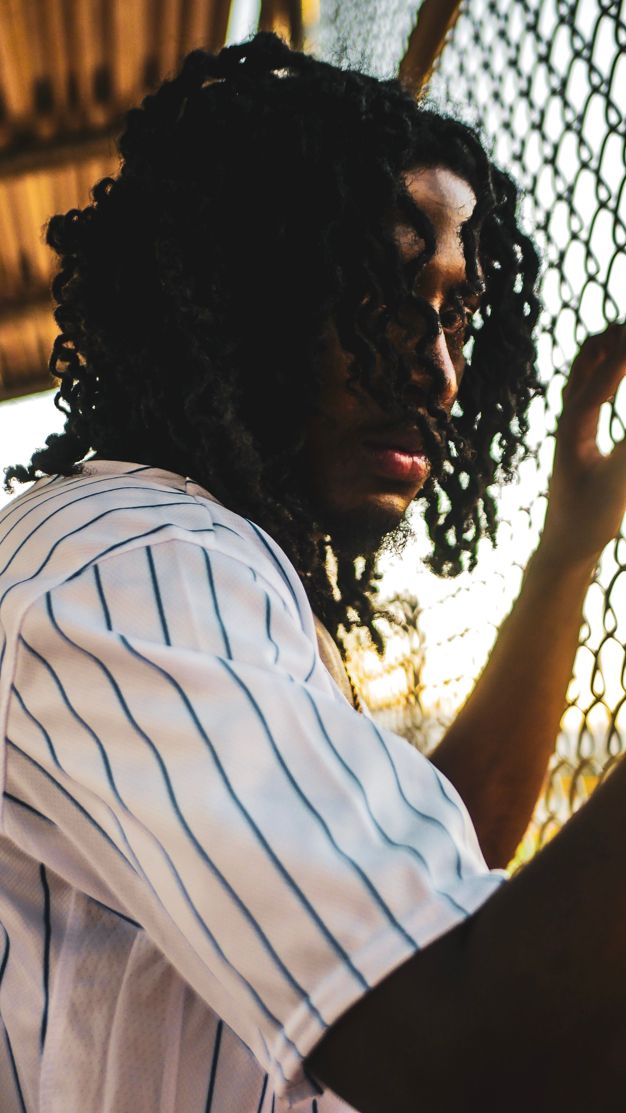 Side view of a serious young adult male with curly hair standing by a metal fence in twilight.