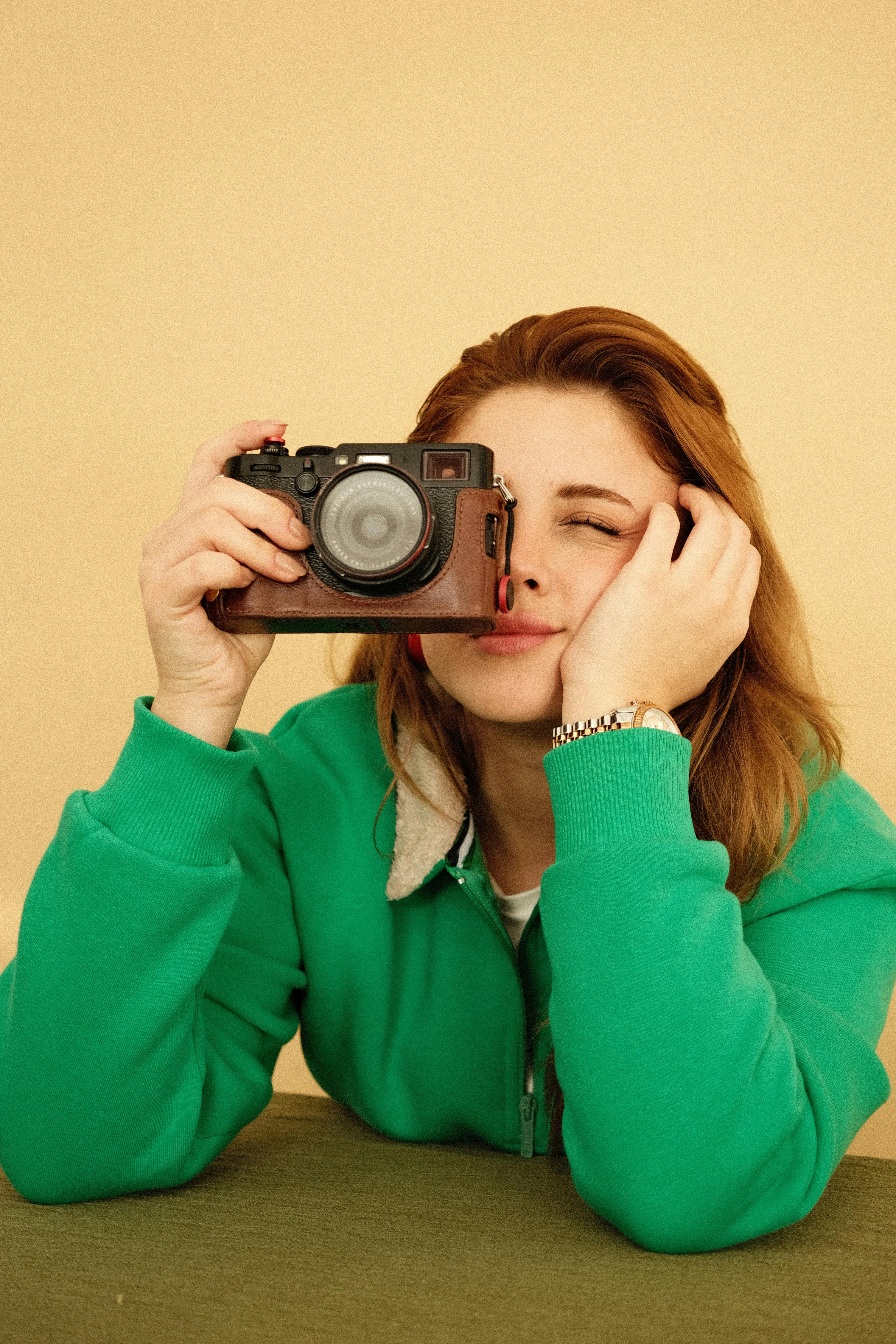 Portrait of a young woman in a green jacket holding a camera indoors.
