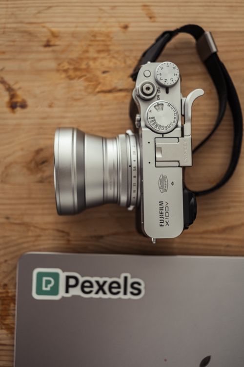 Free A silver camera beside a laptop with a Pexels sticker on a wooden table; ideal for tech-focused imagery. Stock Photo