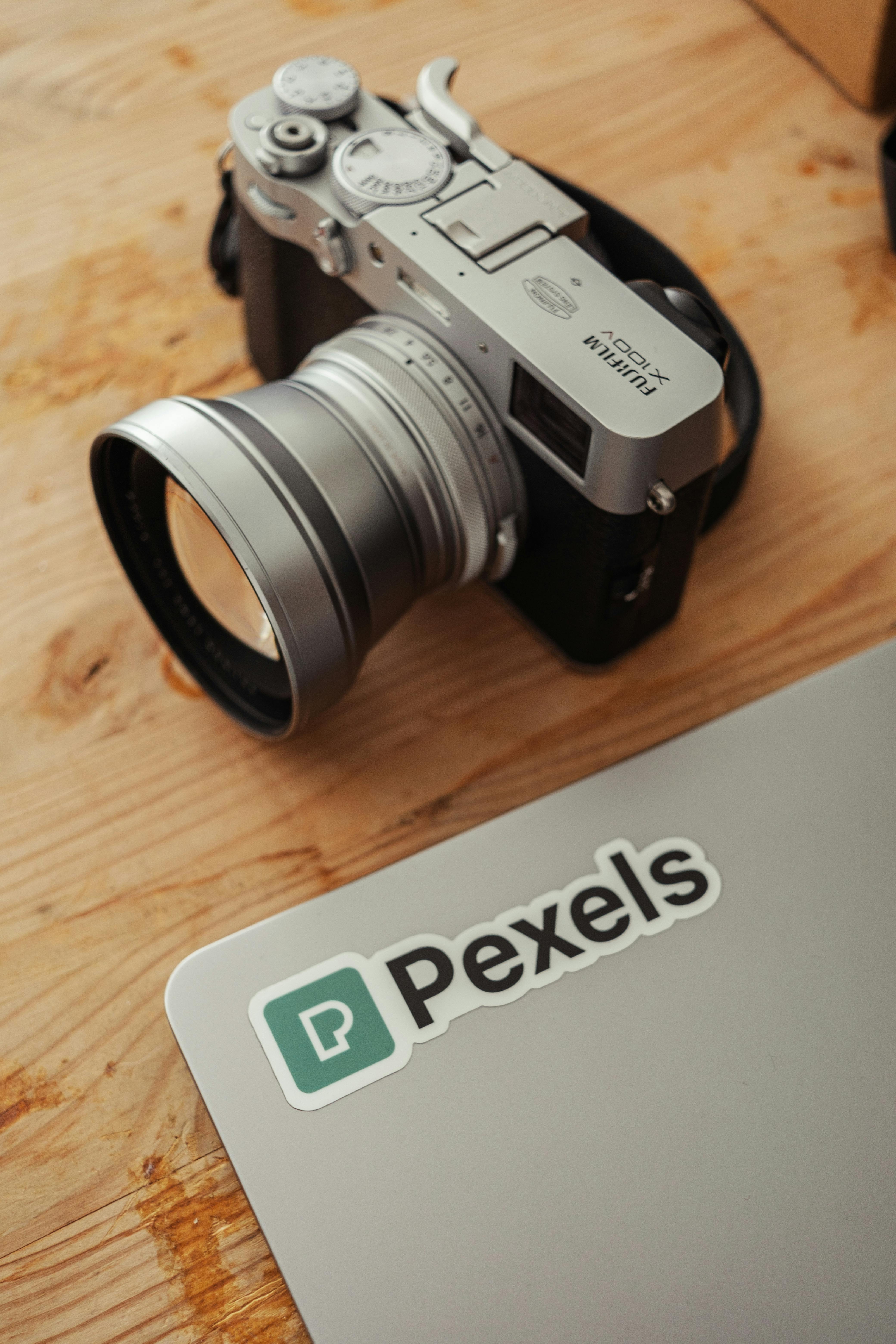 A stylish vintage camera resting on a wooden table alongside a laptop featuring a Pexels sticker.