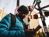 白天，一名黑人男性在有红绿灯和建筑物的城市十字路口使用智能手机