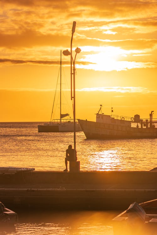 A man stands on a pier at sunset with boats in the background