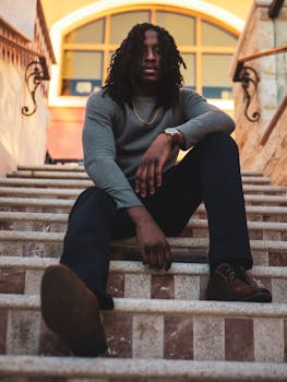 一位时尚男子留着辫子，在户外楼梯上随意摆着姿势，显得十分自信