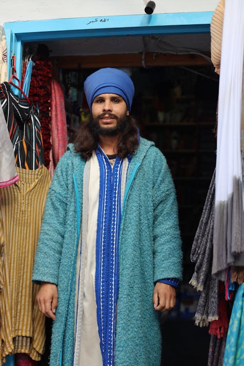 A man in a turban and blue coat stands in front of a shop