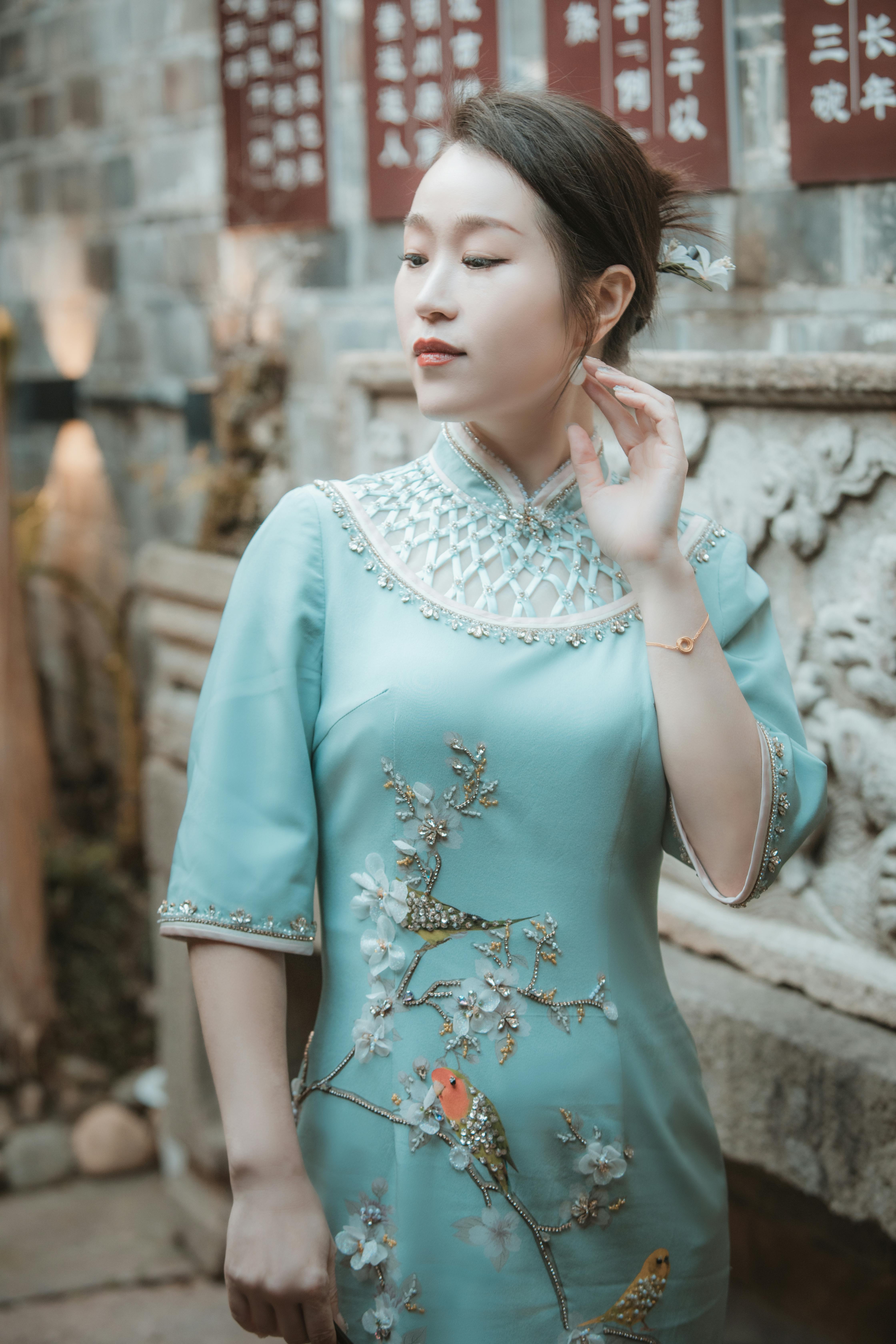 Asian woman posing in elegant traditional dress with an ornate wall backdrop, outdoors.