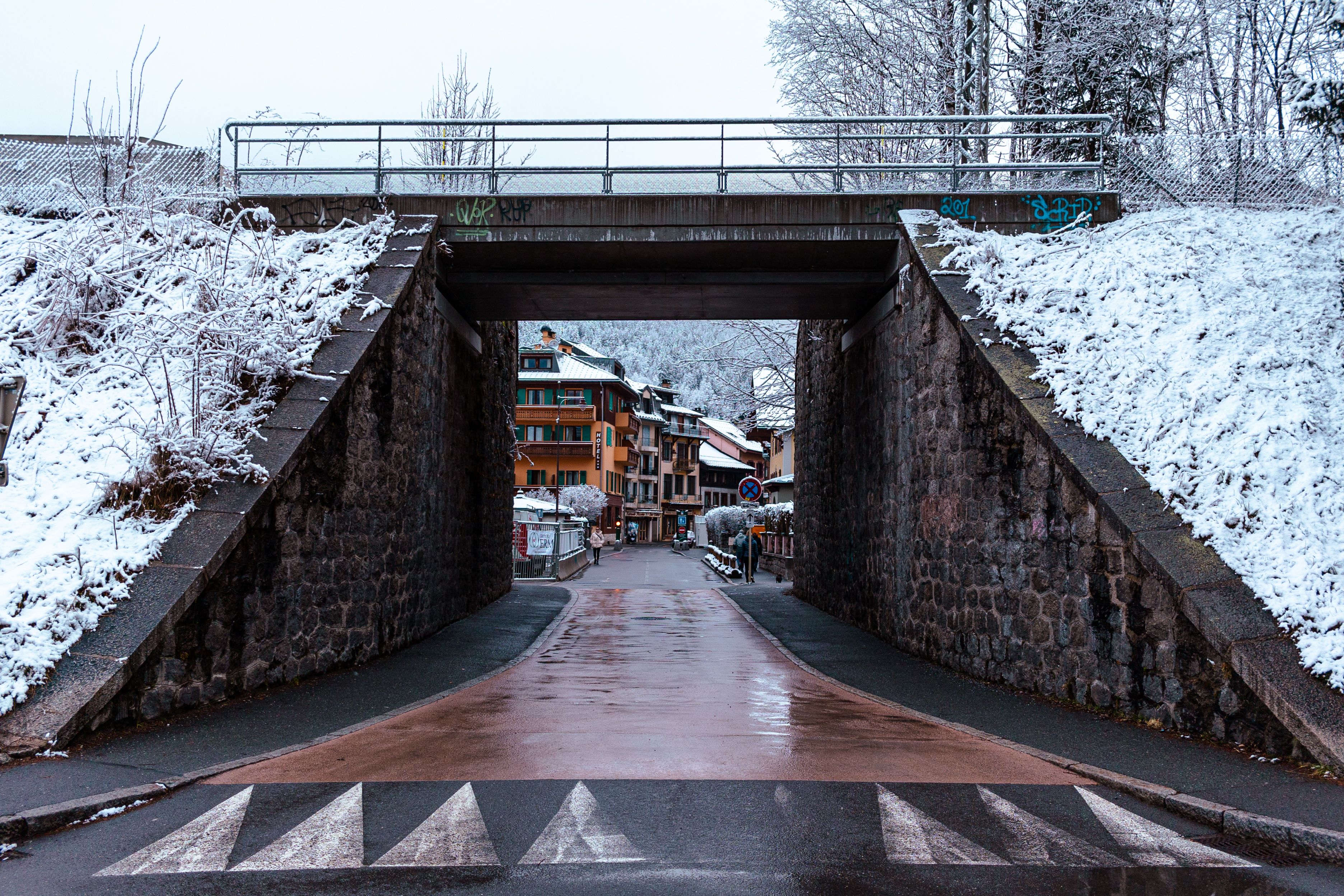 A wet road running under a stone railway bridge into Chamonix, snow banked along both sides and the town's shopfronts beyond