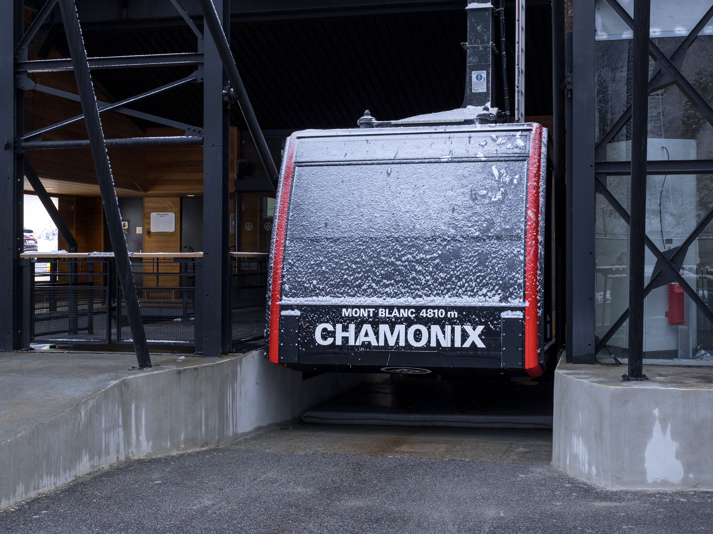 A cable-car cabin with a frosted grey front and red trim, labelled Mont Blanc 4810 m Chamonix, sitting inside a dark steel station building