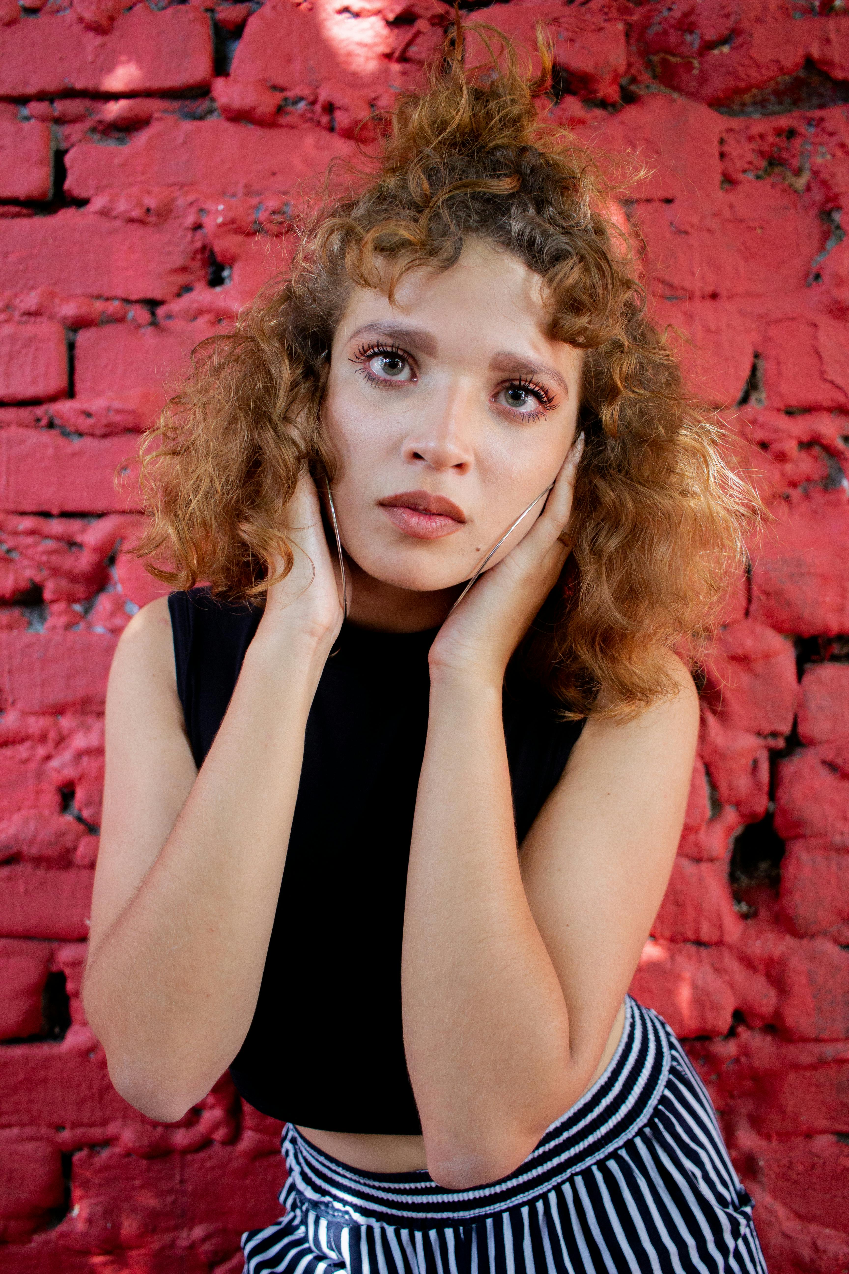 A woman poses with a thoughtful expression against a red brick wall, wearing a striped skirt.