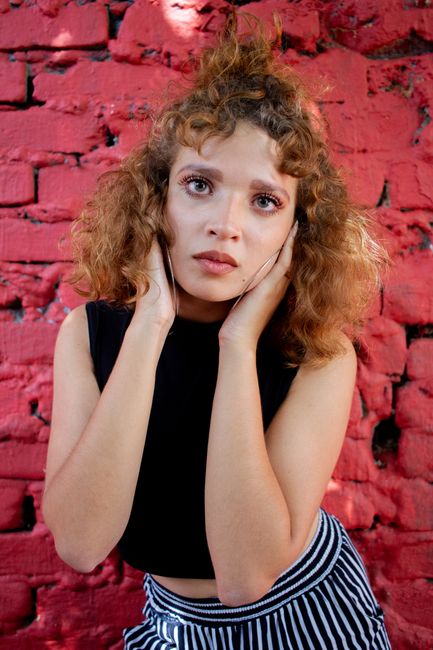 A woman poses with a thoughtful expression against a red brick wall, wearing a striped skirt.