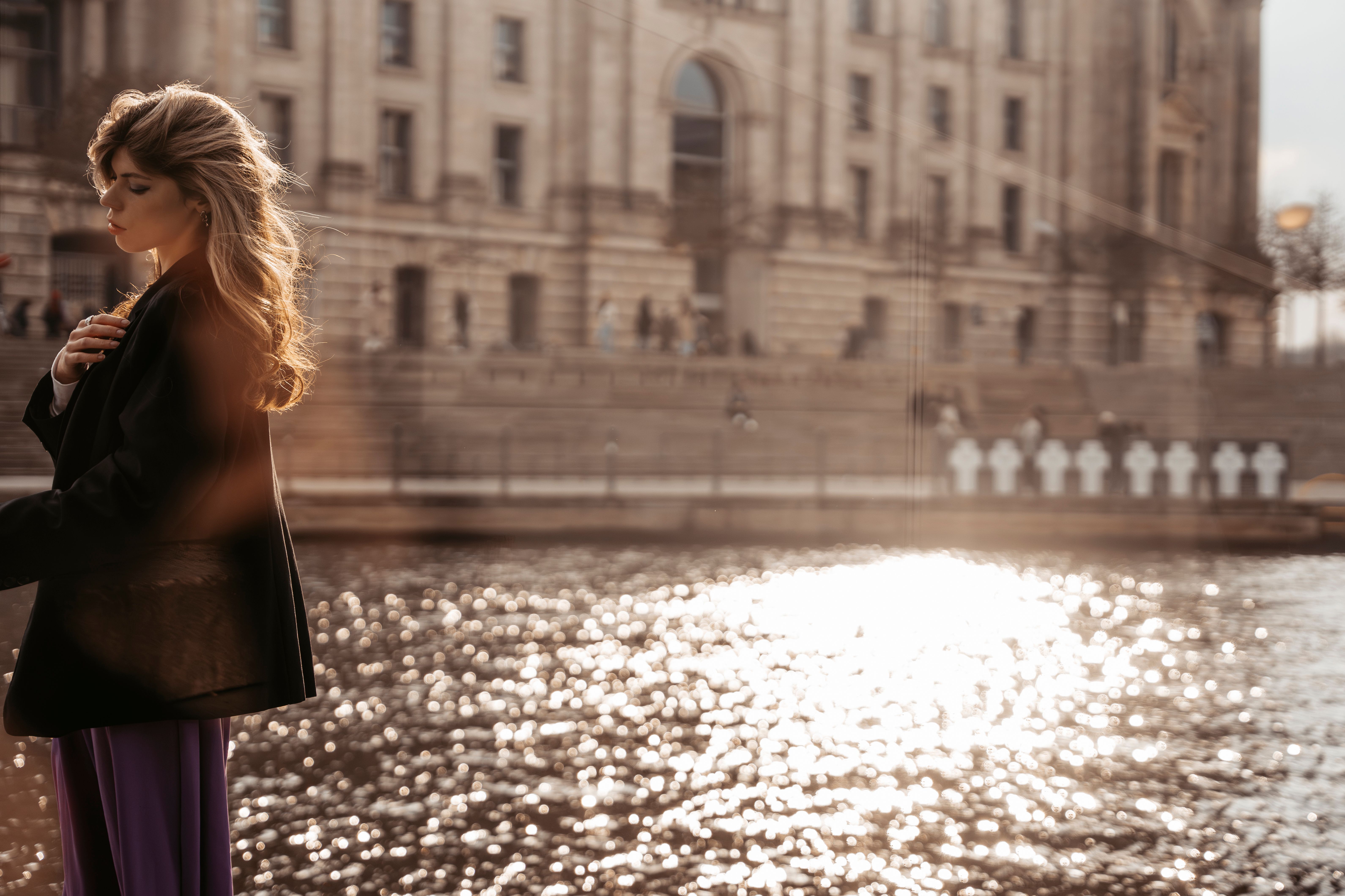 Elegant woman by a sunlit river with urban architecture in the background.