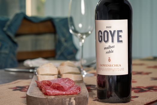 A wine bottle, glass, sliced bread, and salami on a rustic table setting create a cozy dining atmosphere.