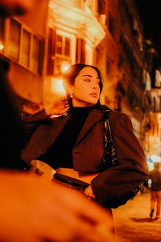 A fashionable woman poses on a city street at night, lit by urban lights.