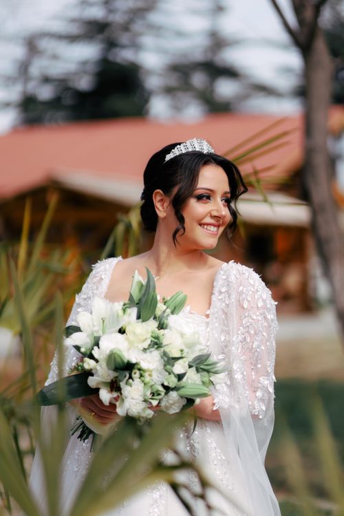 Free A beautiful brunette bride smiles outdoors, holding a white floral bouquet, wearing a tiara and wedding dress. Stock Photo