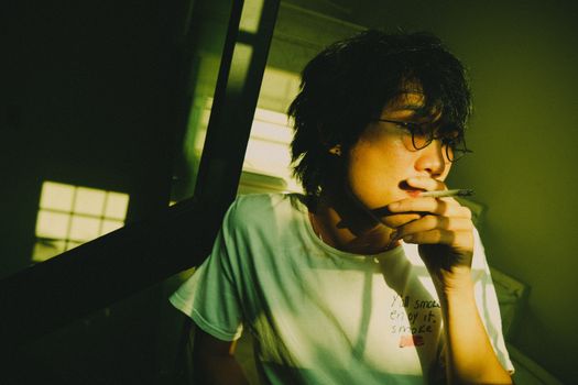 Thoughtful young adult in glasses smoking indoors with dramatic lighting.