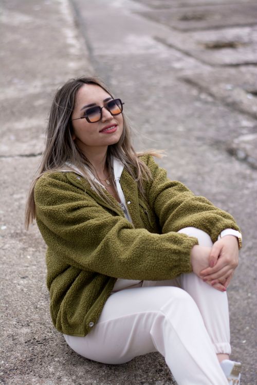 Free Stylish woman in green jacket and white pants sitting outdoors, smiling with sunglasses. Stock Photo