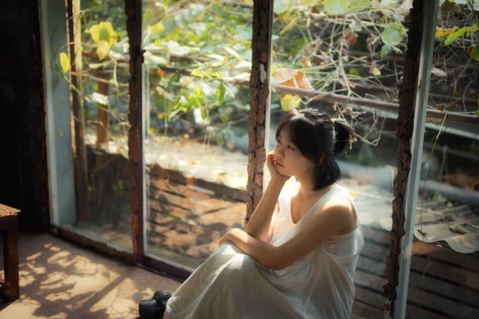 A woman in a white dress sits pensively by a sunlit window surrounded by autumn leaves.