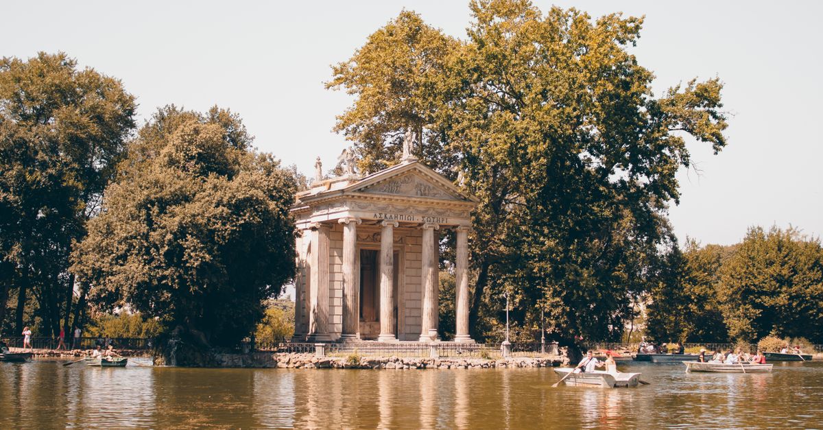 Scenic view of the Temple of Aesculapius surrounded by lush trees and a tranquil lake in Villa Borghese, Rome.