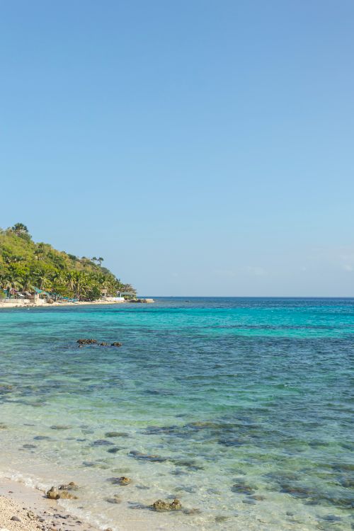 Free Idyllic tropical beach with clear blue waters and lush greenery under a bright sky. Stock Photo
