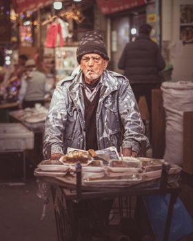 Senior vendor smokes while selling food in a lively street market, creating a moody urban scene.