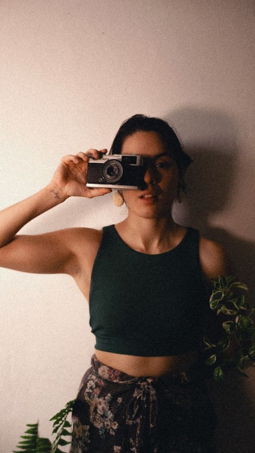 Free Young woman in a black top holding an analogue camera, standing against a white wall with plants. Stock Photo
