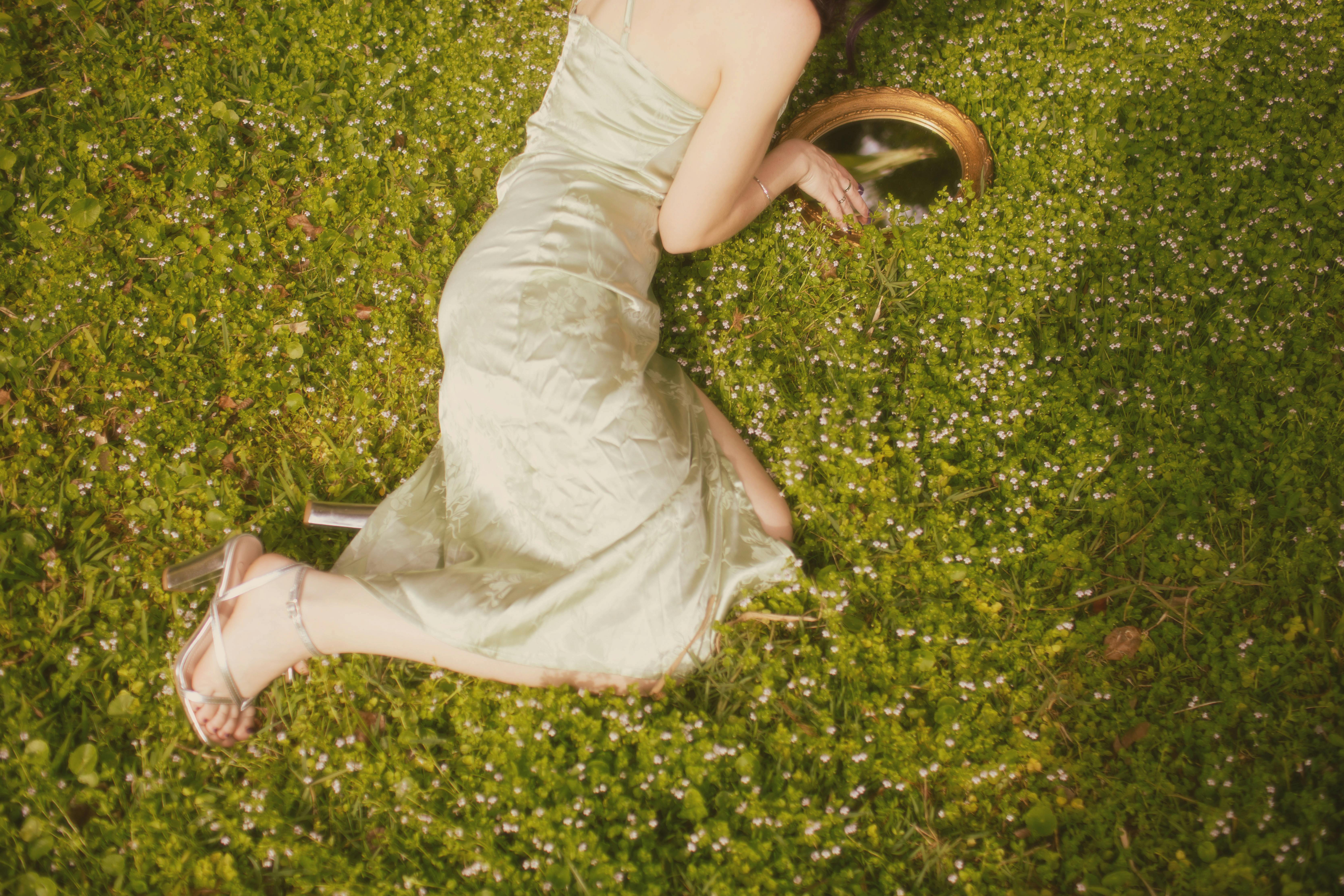 A woman in a light dress lying on a green meadow looking into a vintage mirror.
