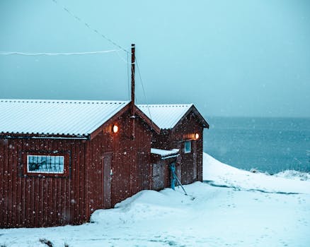 A rustic red cabin during snowfall by the coast in Troms, Norway. Serene winter scene.