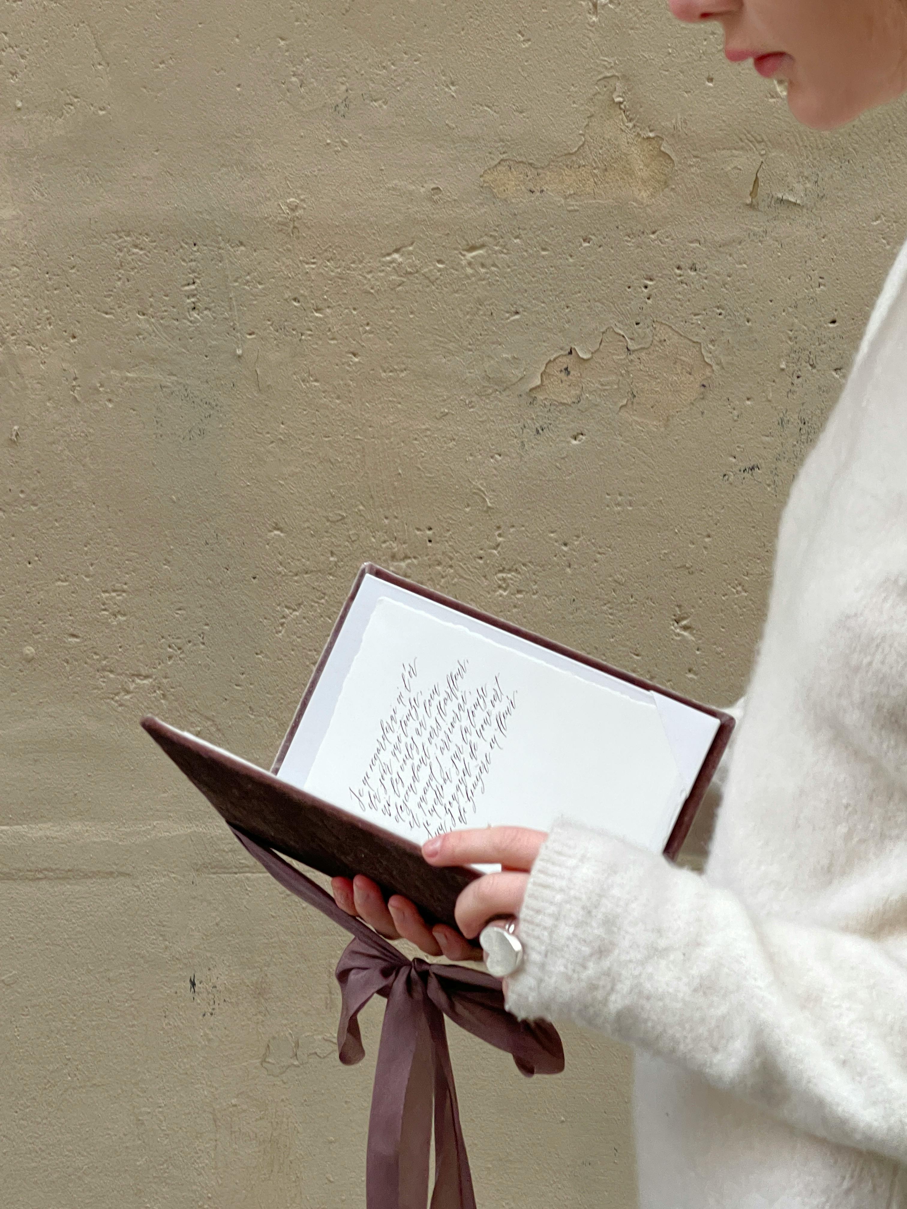 Close-up of person reading a notebook with ribbon, wearing white sweater, against textured wall.