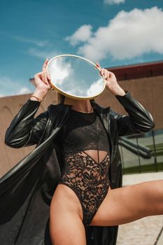Fashion shoot of a woman in lace bodysuit and leather coat holding a mirror outdoors.