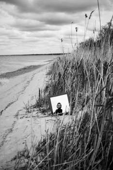 A woman reflected in a mirror on a serene beach surrounded by rushes.
