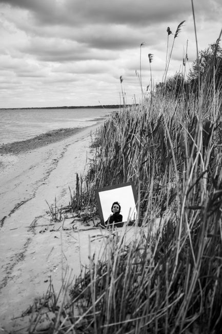 A woman reflected in a mirror on a serene beach surrounded by rushes.