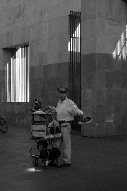 A street vendor with a cart and parrot stands in an urban setting, captured in black and white.