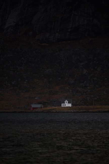 A lone white house and barn by a lake under dark mountain terrain.