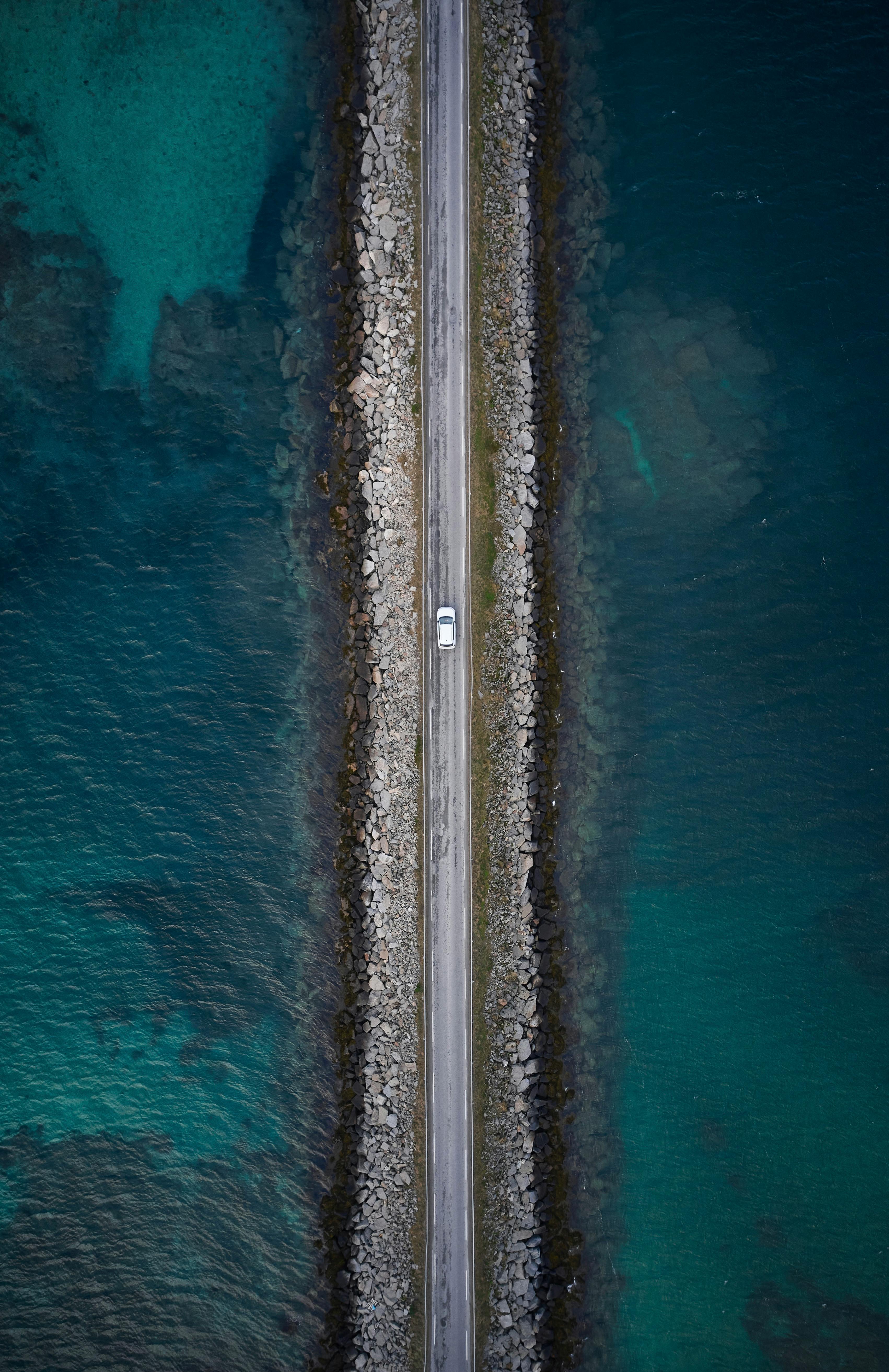 Drone shot capturing a car driving on a narrow coastal road surrounded by water.