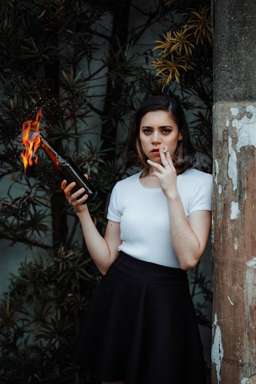 Photo of Woman Leaning on Pole While Holding Molotov Cocktail and Smoking a Cigarette
