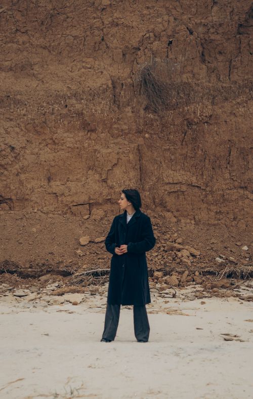 Free Woman in dark coat stands on sandy beach by a tall, earthy cliff. Stock Photo