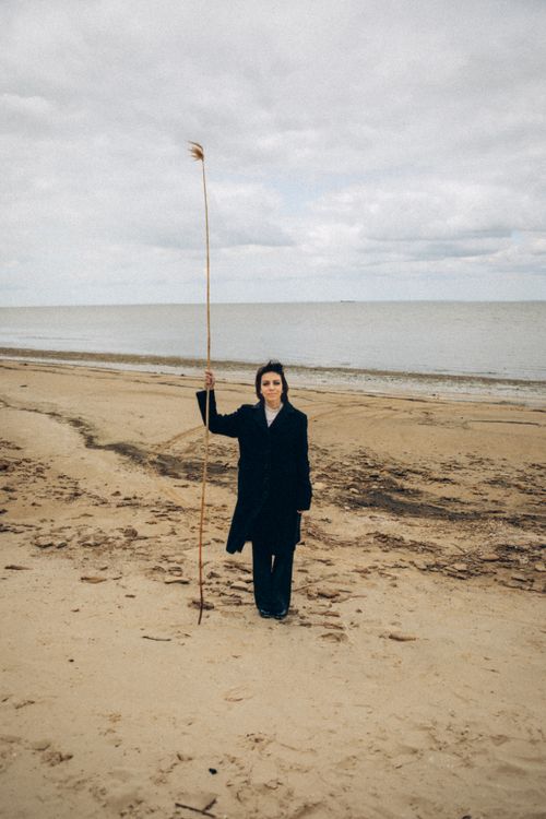 Free Woman in a coat holding a tall stick on a serene sandy beach under a cloudy sky. Stock Photo