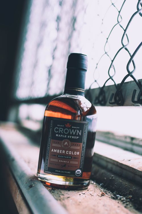 Free A bottle of rich amber Crown maple syrup positioned beside a rustic window, radiating warmth. Stock Photo