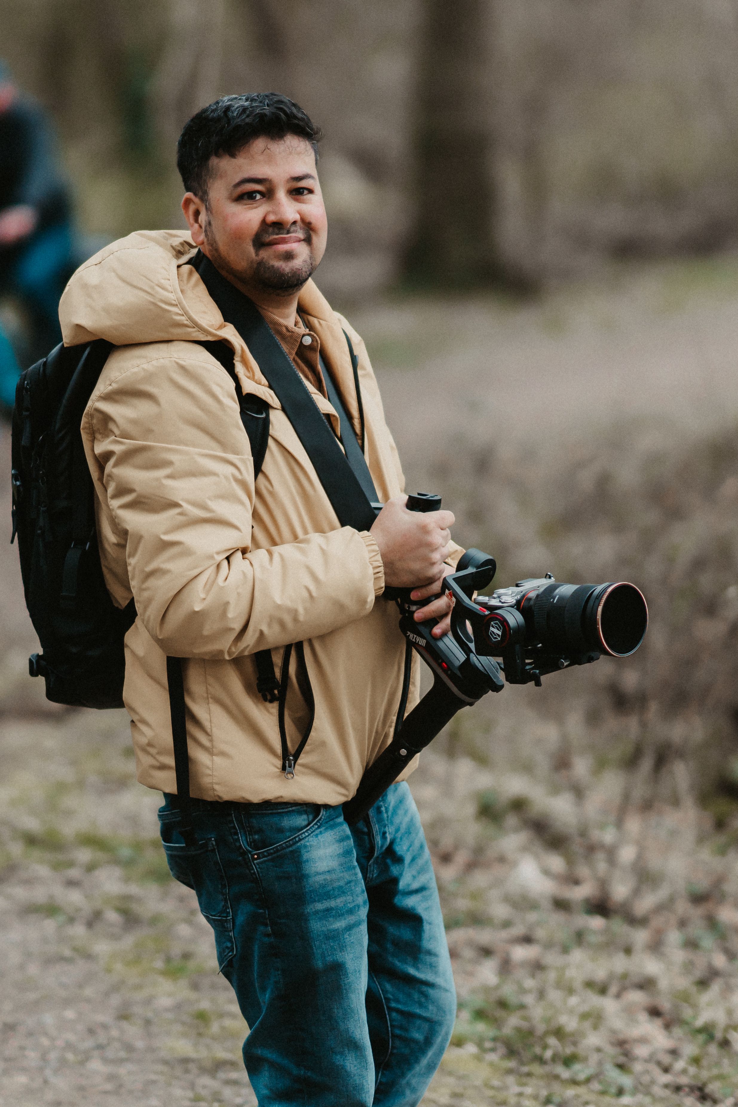 Man in beige jacket with camera smiling, outdoors in a natural setting.