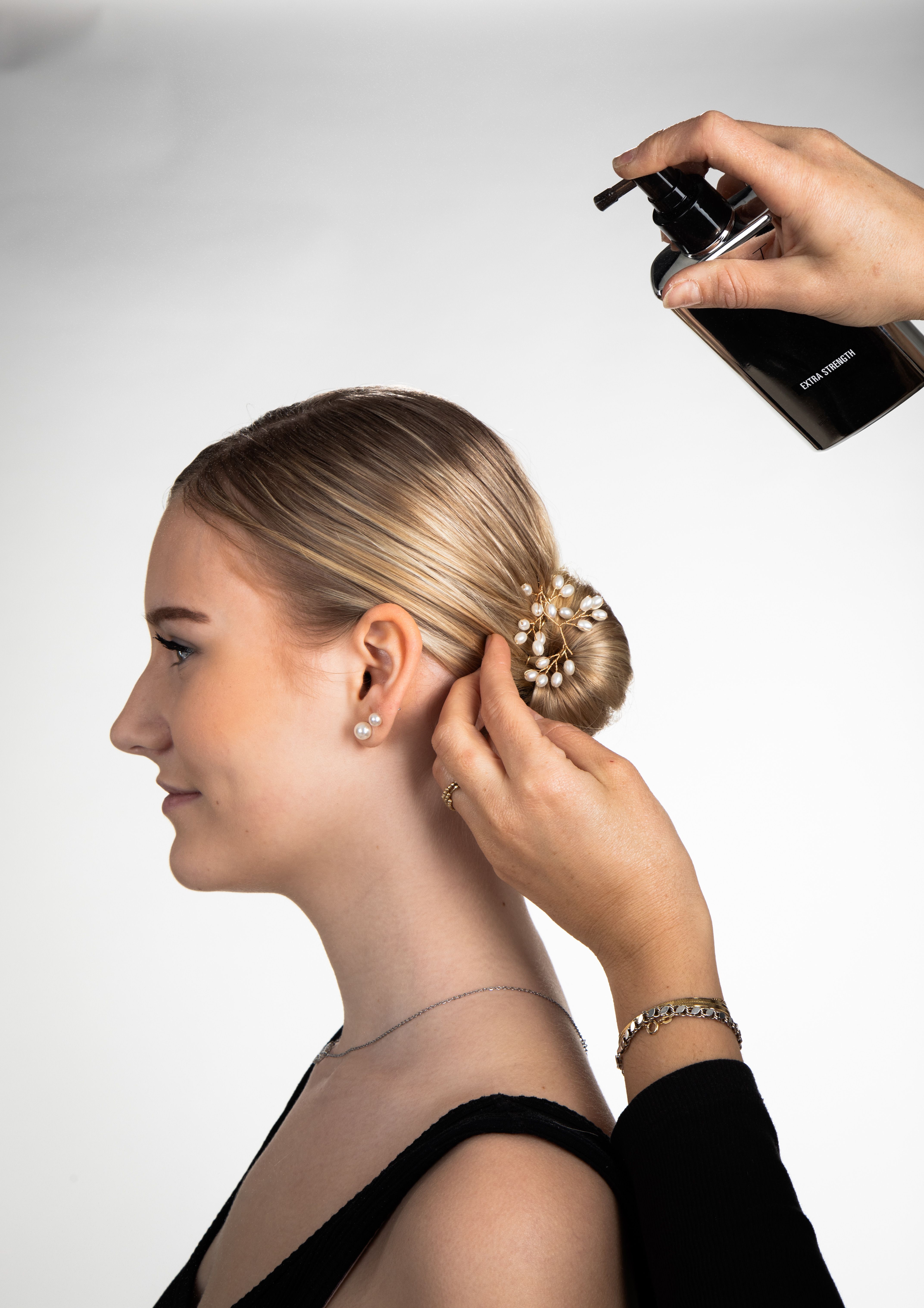Professional hairstylist applying product to elegant bun on woman with pearl accessory.
