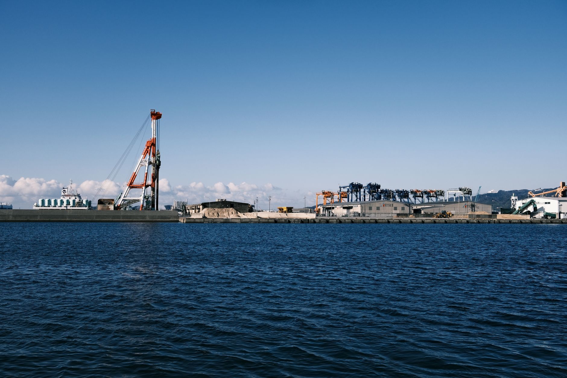 View of an industrial port in Gamagōri, Japan, showcasing cranes and the coastal area under a clear sky.