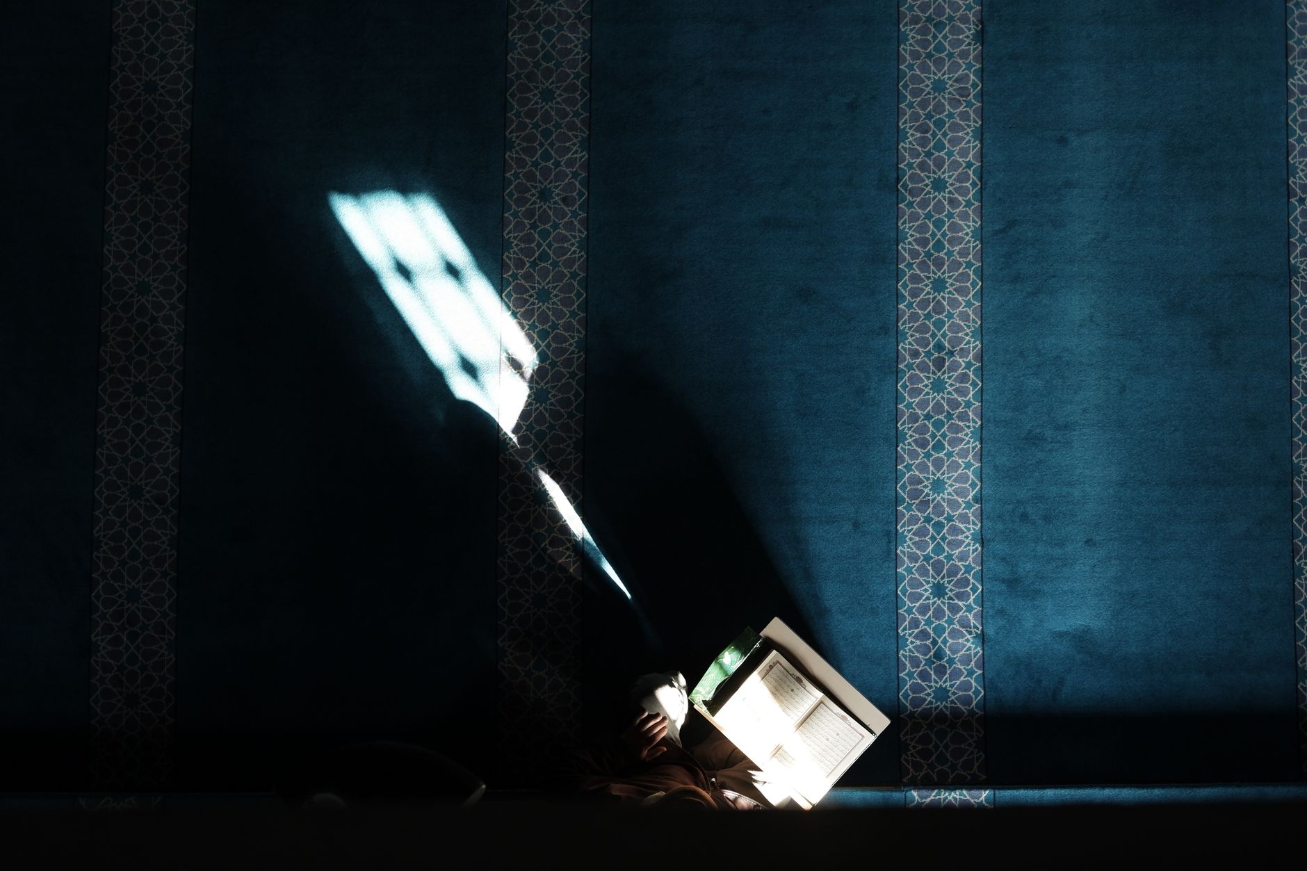 A student reading the Quran in a mosque, illuminated by sunlight through a window