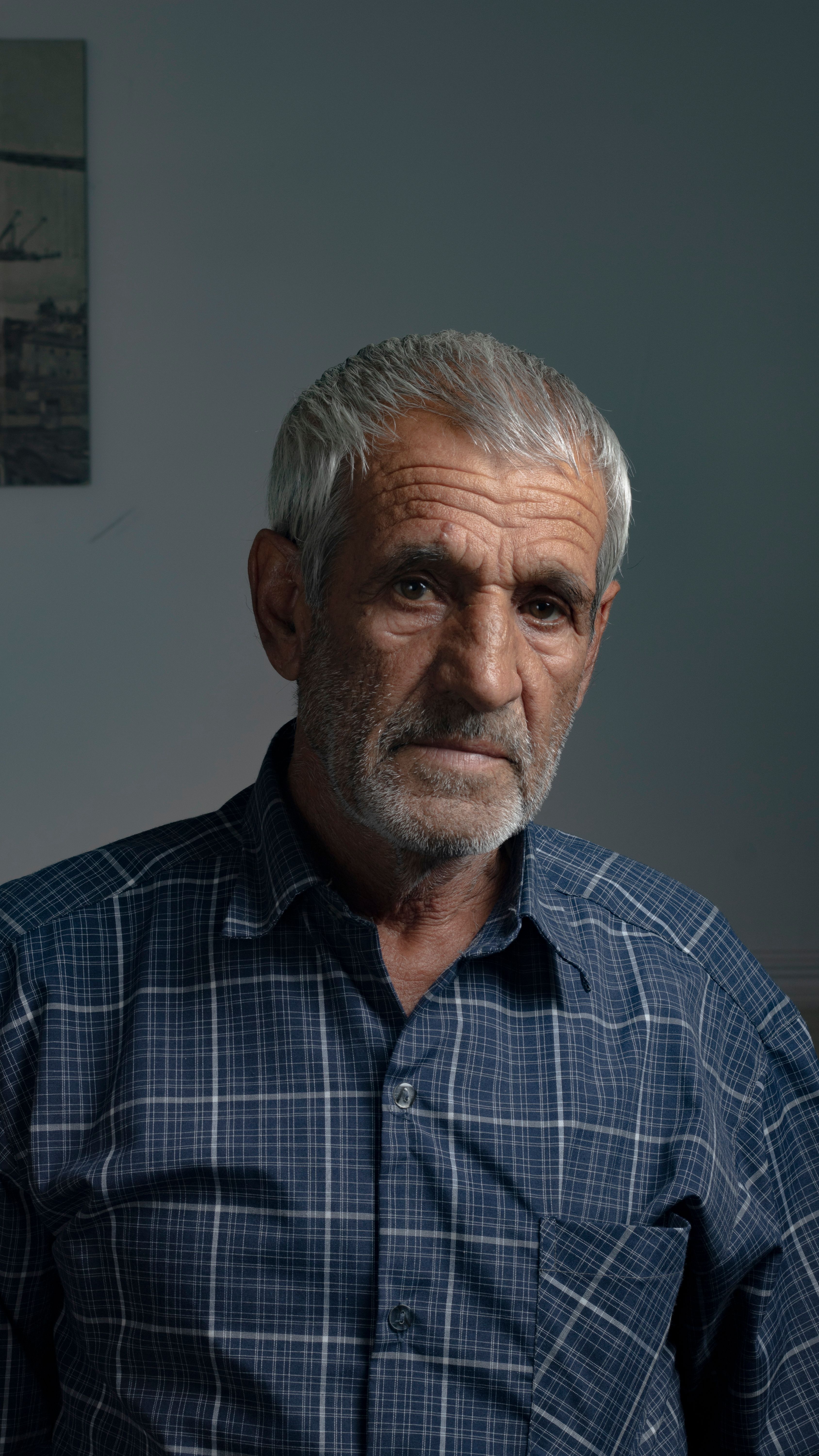 Elderly man with gray hair in a blue checkered shirt against a gray background.
