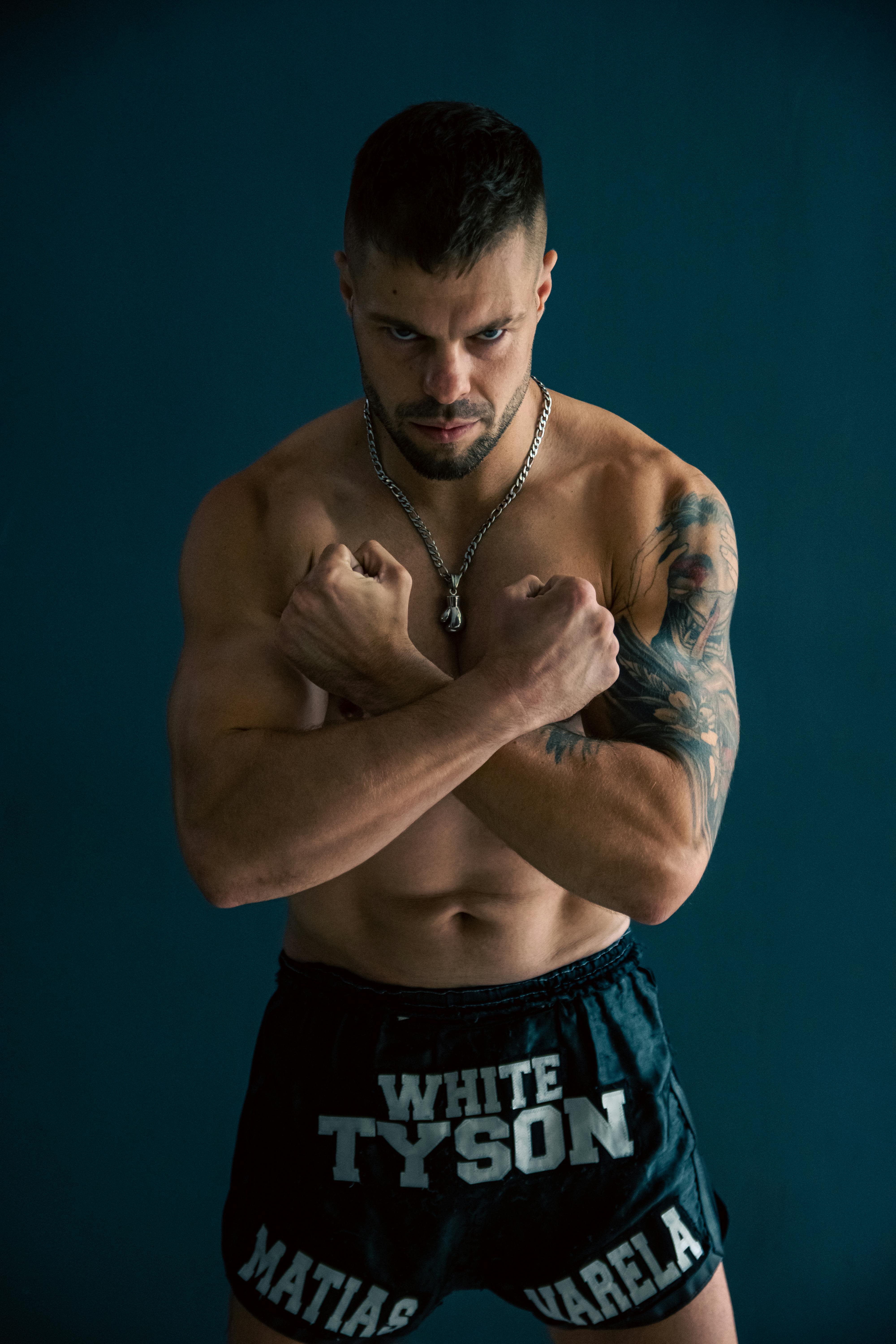 Athletic man posing shirtless showing tattoos and muscles, on a blue background.