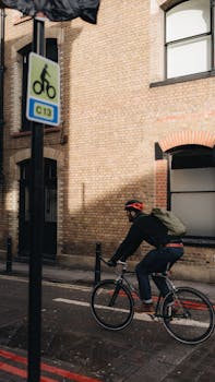 A man cycling with a backpack on an urban street, passing a brick building.
