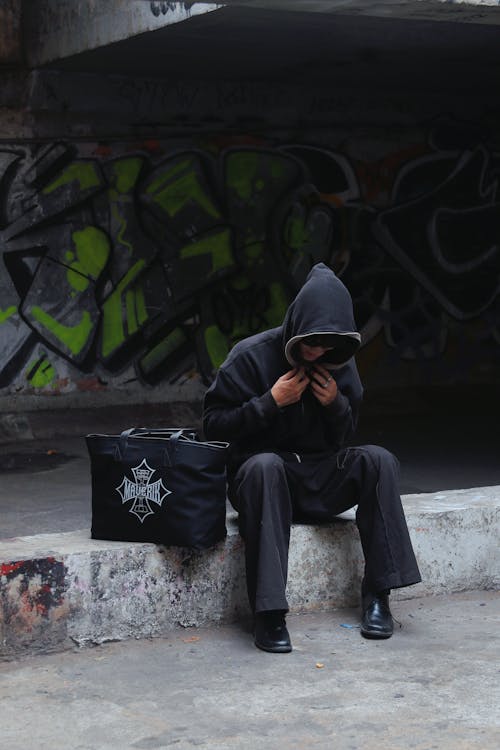 Man in Black Hoodie Sitting with Bag on Wall