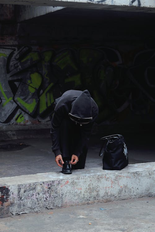 A person in a hoodie is kneeling down in front of a graffiti wall
