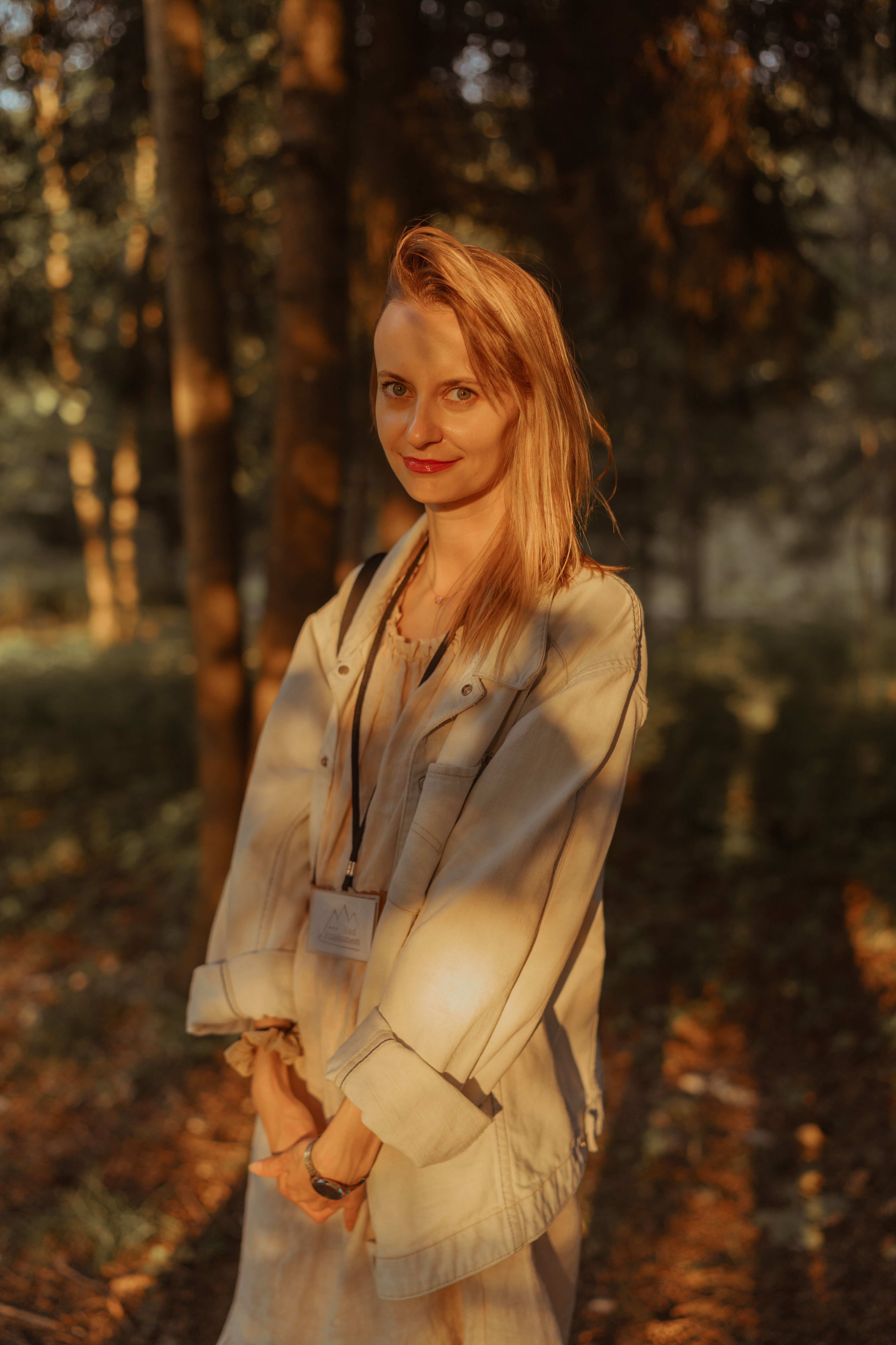 A woman with blonde hair stands in a sunlit forest, wearing a light jacket.